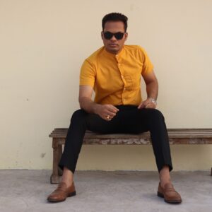 Fashionable man in yellow shirt and sunglasses sitting on bench outdoors.